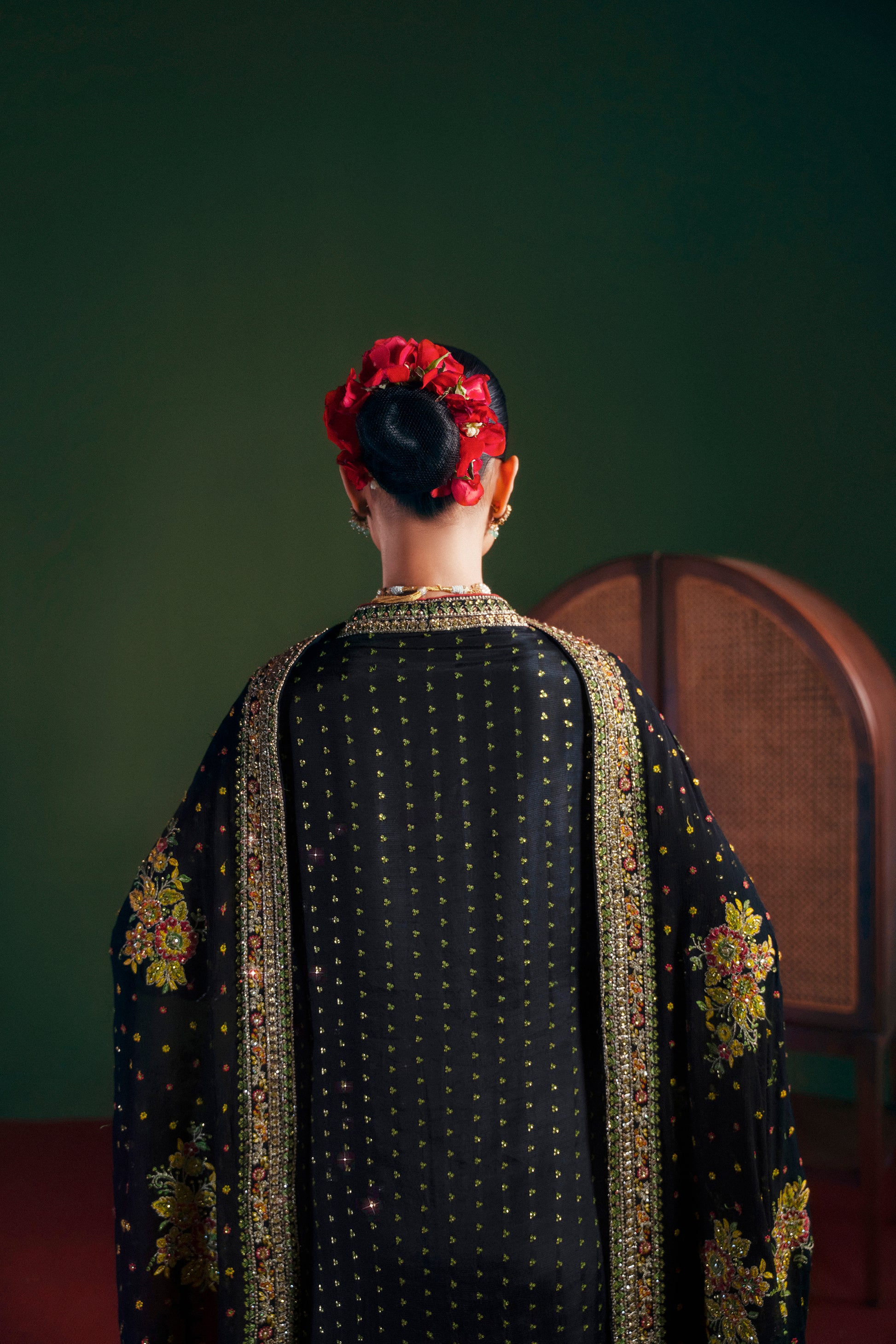 Person wearing a black embroidered garment with gold details, sitting on a wooden chair against a dark green wall| Multicolor embroidered PK silk long shirt close-up